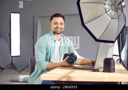 Ein junger, glücklicher Fotograf, der am Schreibtisch sitzt, mit Kamera und Linsen und lächelt. Stockfoto