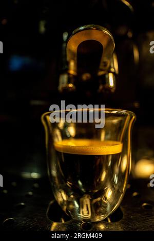 Makroansicht im goldenen Licht einer Glasbecher mit modernem Design und frisch gebrühtem schwarzen Kaffee mit goldenem Schaum und kleinen Luftblasen auf einem Barista Automat Stockfoto