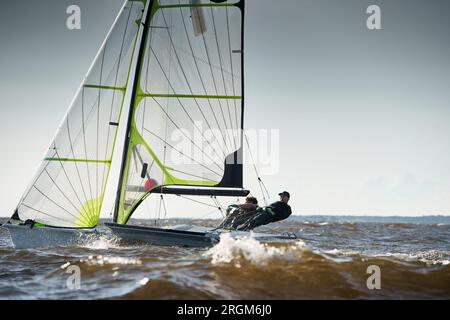 Russland, St. Petersburg, 07. Juli 2023: Die Aussicht auf das Sportsegelboot ist bei Sonnenuntergang hoch, Sportler neigen das Boot, Geschwindigkeit und Spritzwasser, Reflexion der Sonne Stockfoto