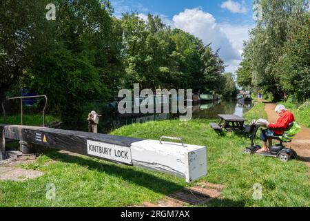 Kintbury Lock am Kennet und Avon Canal in Berkshire, England, Großbritannien, mit einer alten Seniorin, die auf einem Roller sitzt und eine Zeitung liest Stockfoto