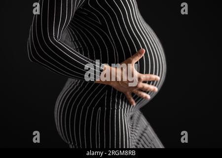 Schwangere Frau, die Hände auf dem Bauch in gestreiftem Kleid mit schwarzem Hintergrund hält Stockfoto