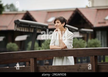 Pensive ältere Frau, die auf einer Holzbrücke steht und wegschaut, altes Leben, verschwommener Hintergrund Stockfoto