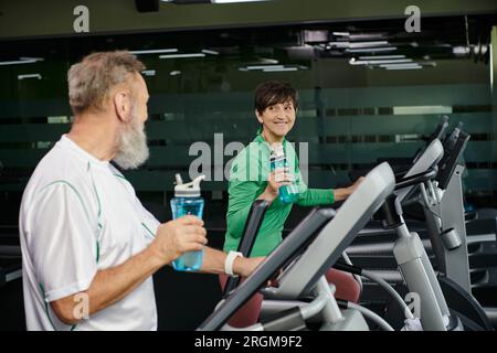 Eine fröhliche Frau, die einen älteren Mann, Mann und Frau ansieht, die im Fitnessstudio trainieren und Sportflaschen in der Hand halten Stockfoto