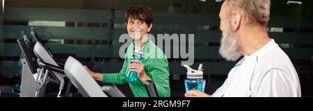Glückliche Frau, die einen älteren Mann, Mann und Frau im Fitnessstudio ansieht, Sportflaschen in der Hand hält, Banner Stockfoto