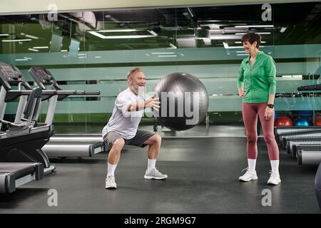 Aufgeregter Seniorenmann, der Squats mit Fitnessball in der Nähe von Frau, Mann und Frau im Fitnessstudio macht, Paar, Sport Stockfoto