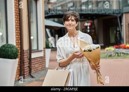 Alternde Bevölkerung, zufriedene Seniorin mit Einkaufstaschen und Blumenstrauß in der Nähe des Outlets, draußen Stockfoto