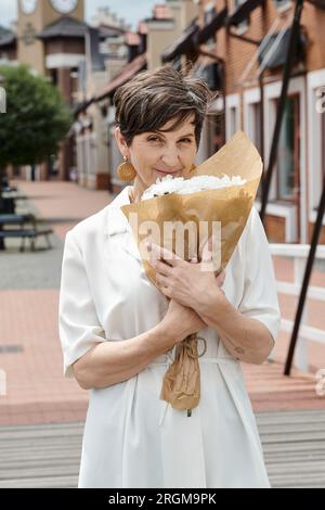 Ältere Frau, die einen Blumenstrauß in der Hand hält und in die Kamera schaut, urbane Kulisse, Sommer, weißes Outfit Stockfoto