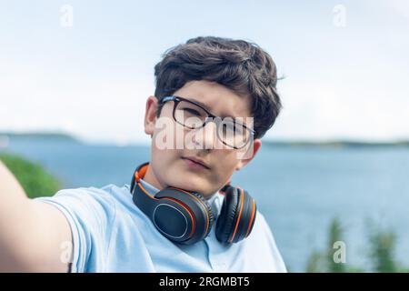 Porträt eines 11-jährigen Jungen mit Brille und kabellosen Kopfhörern, Selfie, Meer im Hintergrund Stockfoto