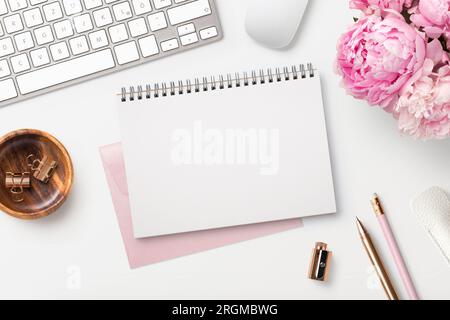 Femininer Arbeitsplatz/Schreibtisch mit offenem Notizblock, Tastatur, stilvollem Büro-/Schreibzubehör und pinkfarbenen Pfingstrosen auf weißem Hintergrund, Draufsicht Stockfoto