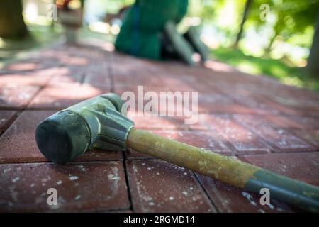 Auf den Pflastersteinen liegt ein Hammer mit Gummispitzen, und im Hintergrund legt ein Arbeiter den Bürgersteig. Stockfoto