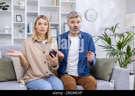 Portrait von enttäuschter betrogener Familie, Mann und Frau, reifes Paar, das traurig auf dem Sofa sitzt und in die Kamera schaut, mit Kreditkarte und Telefon der Bank online einkaufen im Internet. Stockfoto