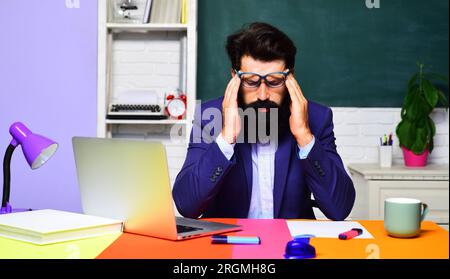 Müder, gestresster Lehrer mit Kopfschmerzen, der im Klassenzimmer am Schreibtisch sitzt. Universitätsprofessor denkt über Probleme nach. Erschöpfter männlicher Schüler Stockfoto