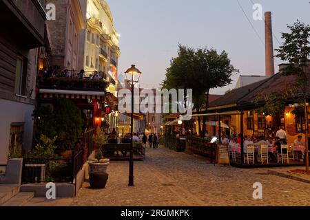 Die böhmische Kopfsteinpflasterstraße Skadarlija mit Leuten in Cafés und Restaurants. Schornstein der alten Brauerei rechts. Belgrad, Serbien. August 2023. Stockfoto