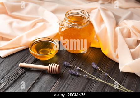 Honig in Glasgefäß und Schüssel, Holzdipper, Lavendelblumen auf dunklem Holz und hellem Textilhintergrund. Der Begriff der Bienenprodukte. Stockfoto