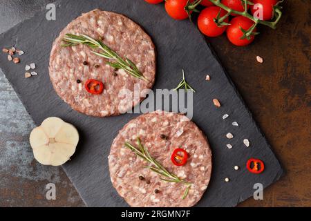 Rohe, frisch gemahlene Rinderburger, Rosmarin, Gemüse und Gewürze auf einer schwarzen Steinplatte auf dunklem Hintergrund, Draufsicht. Fleisch halbfertig Stockfoto