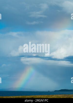 Langer Regenbogenstrahl, der durch die Wolkenschichten und den sonnenbeleuchteten blauen Himmel über dem Meer schneidet und eine Dreiecksform mit einer Wolke schafft Stockfoto