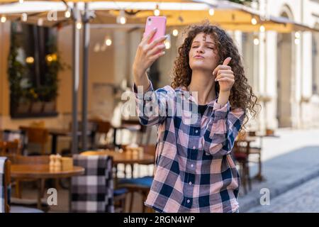 Hübsche junge Frau Bloggerin macht Selfie auf Smartphone, kommuniziert Videoanrufe online mit Abonnenten, nimmt Geschichten für soziale Medien Vlog im Freien auf. Ein Mädchen, das auf der sonnigen Stadtstraße spaziert Stockfoto