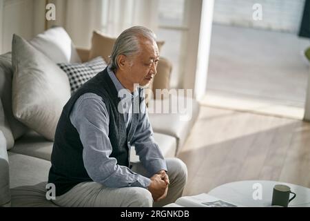 Trauriger asiatischer Seniorenmann, der zu Hause auf der Couch im Wohnzimmer sitzt Stockfoto