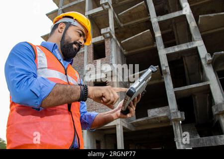 Junger indischer Bauingenieur oder Architekt mit Helm und Weste und digitalem Tablet-Blueprint auf der Baustelle. Niedriger Winkel. Stockfoto