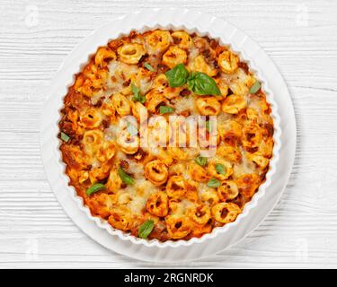 Tortellini gebacken mit Mozzarella-Käse, Tomatensoße und Rinderhack in weißer Backform auf weißem Holztisch, horizontale Sicht von oben, flach liegend Stockfoto