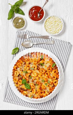 Tortellini gebacken mit Mozzarella-Käse, Tomatensoße und Rinderhack in weißer Backform auf weißem Holztisch mit Löffel und Gabel, vertikaler Blick fr Stockfoto