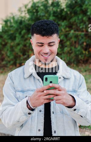 Vertikales Porträt eines lächelnden marokkaners, der Spaß mit einem Smartphone hat, um ein Videospiel zu spielen. Glücklicher Teenager, der mit einem Handy Nachrichten in den sozialen Medien schickt. Junger Mann mit einem Handy. Hochwertiges Foto Stockfoto