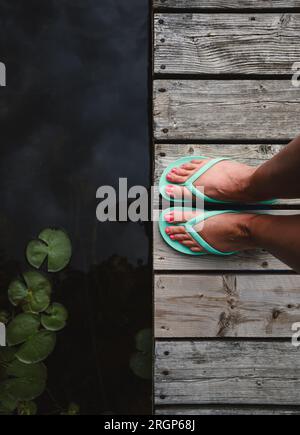 Über den Füßen der Frau in Sandalen auf einem abgenutzten Holzdock am See. Stockfoto