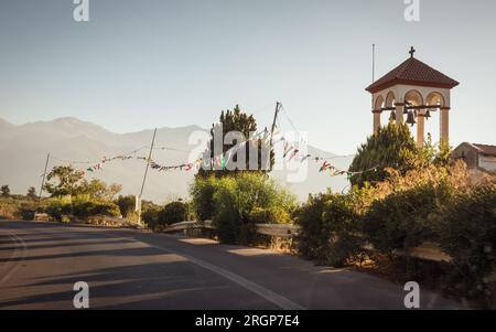 Fahrt im Norden Kretas. Kleine Kapelle am Straßenrand bei Sonnenuntergang mit Bergsilhouetten im Hintergrund. Lange Schatten von der untergehenden Sonne auf der rechten Seite. Stockfoto