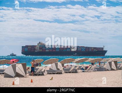 MIAMI, FL, USA - 22. FEBRUAR 2023: Menschen, die sich am Miami South Beach entspannen, großes Containerschiff, das im Hintergrund vorbeifährt. Stockfoto