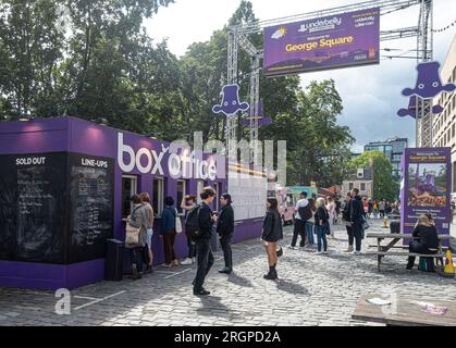 Randgänger, die Tickets am Underbelly Box Office in George Square, Edinburgh kaufen, während andere auf die Aufzählung schauen, Schlange für Essen an der Strasse Stockfoto