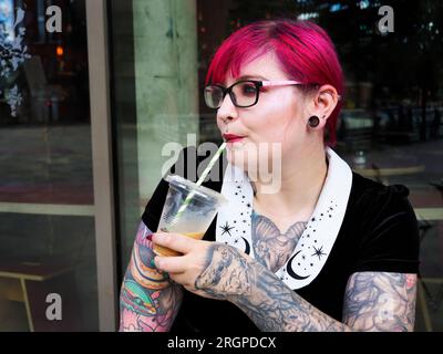 Eine Frau Mitte zwanzig mit gefärbtem rotem Haar und Tätowierungen, die Gläser trägt und vor einem Café in Leeds West Yorkshire, England, Eiskaffee trinkt Stockfoto