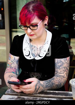 Eine Frau Mitte zwanzig mit gefärbtem rotem Haar und Tätowierungen, die eine Brille mit einem Smartphone trägt, in Leeds West Yorkshire, England Stockfoto