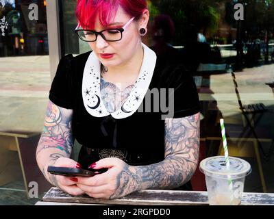 Eine Frau Mitte zwanzig mit gefärbtem rotem Haar und Tätowierungen, die eine Brille mit einem Smartphone trägt, in Leeds West Yorkshire, England Stockfoto
