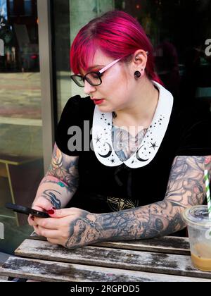 Eine Frau Mitte zwanzig mit gefärbtem rotem Haar und Tätowierungen, die eine Brille mit einem Smartphone trägt, in Leeds West Yorkshire, England Stockfoto