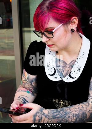 Eine Frau Mitte zwanzig mit gefärbtem rotem Haar und Tätowierungen, die eine Brille mit einem Smartphone trägt, in Leeds West Yorkshire, England Stockfoto