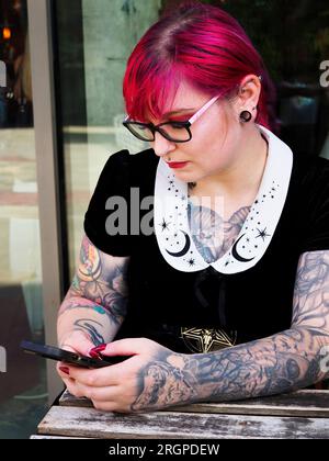 Eine Frau Mitte zwanzig mit gefärbtem rotem Haar und Tätowierungen, die eine Brille mit einem Smartphone trägt, in Leeds West Yorkshire, England Stockfoto