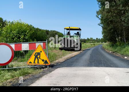 Warnschild für Straßenbauarbeiten an einem Zaun einer neuen Baustelle. Fertigerfertiger in bg. Raupenpflastermaschine zum Verlegen von Asphalt. Stockfoto