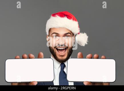 Ein glücklicher, aufgeregter Geschäftsmann in Santa hat, der zwei Smartphones zeigt. Smartphone mit leerem weißen Display für neue Anwendung oder mobiles Websit Stockfoto