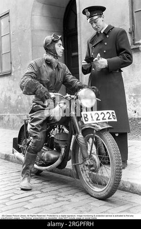 Motorradfahrer in den 1950er. Ein junger Mann, gekleidet wie die Motorradfahrer in den 1950er Jahren. Ganz aus Leder. Lederstiefel, -Hose und -Jacke. Ein Nierengurt außerhalb der Jacke. Aus irgendeinem Grund steht ein Polizist an seiner Seite auf dem Bürgersteig und schaut sich den Motorradfahrer-Führerschein an. Schweden 1955. Er ist Schauspieler Sven-Eric Gamble, 1924-1976. Kristoffersson Ref. 2B Stockfoto