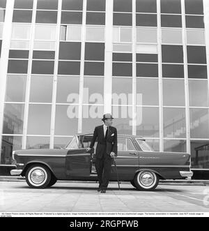 In den 1960er Jahren. Ein Mann steht vor seinem Auto, einem Opel Kapitän, vor einem Hochhaus mit Glasfenstern, die einen zerdrückten Hintergrund bilden. Er ist angezogen wie ein Geschäftsmann in Anzug und Hut, hält einen Regenschirm und eine Aktentasche in jeder Hand. Schweden 1962. Kristoffersson Ref. C98-4 Stockfoto