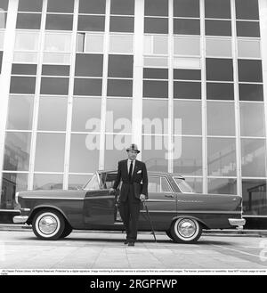 In den 1960er Jahren. Ein Mann steht vor seinem Auto, einem Opel Kapitän, vor einem Hochhaus mit Glasfenstern, die einen zerdrückten Hintergrund bilden. Er ist angezogen wie ein Geschäftsmann in Anzug und Hut, hält einen Regenschirm und eine Aktentasche in jeder Hand. Schweden 1962. Kristoffersson Ref. CV9-11 Stockfoto