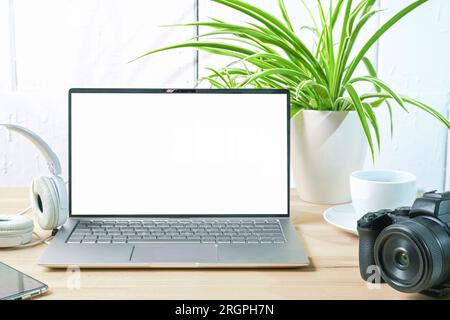 Offener Laptop mit einem leeren weißen Bildschirm, Kamera, Kopfhörern und einer Pflanze auf dem Schreibtisch eines Journalisten oder Fotografen an einer weiß bemalten Wand, Mock-U Stockfoto