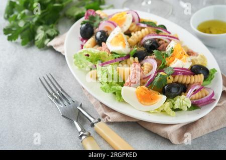 Thunfischsalat mit Pasta, Eiern, Kartoffeln, Oliven, roten Zwiebeln und Sauce auf weißem Teller auf altem hellgrauem Betontisch. Nicoise-Salat. Franzosen Stockfoto
