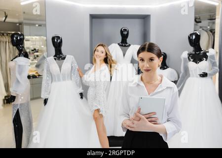 Attraktive Ladenbesitzer für Brautläden verwenden ein digitales Tablet, um Kunden bei der Auswahl des Hochzeitskleids im Geschäft zu helfen. Stockfoto