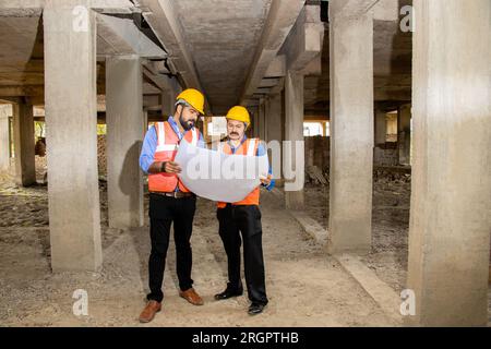 Zwei indische Bauingenieure oder Architekten, die Helm und Weste tragen, mit Papierkram auf der Baustelle, um Immobilienprojekte zu besprechen, Stockfoto