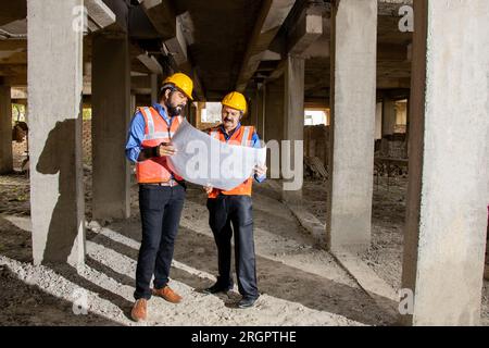 Zwei indische Bauingenieure oder Architekten, die Helm und Weste tragen, mit Papierkram auf der Baustelle, um Immobilienprojekte zu besprechen, Stockfoto