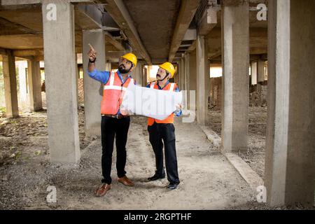 Zwei indische Bauingenieure oder Architekten, die Helm und Weste tragen, mit Papierkram auf der Baustelle, um Immobilienprojekte zu besprechen, Stockfoto