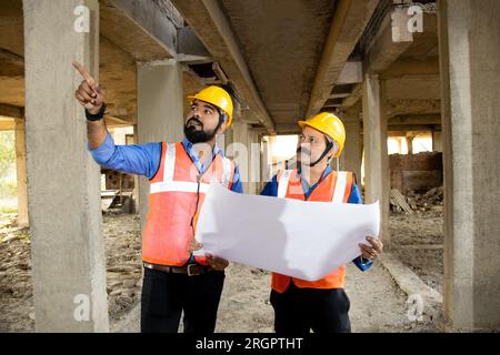 Zwei indische Bauingenieure oder Architekten, die Helm und Weste tragen, mit Papierkram auf der Baustelle, um Immobilienprojekte zu besprechen, Stockfoto