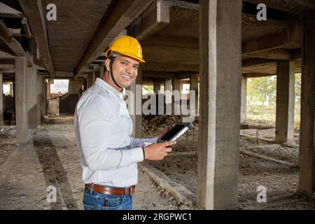 Porträt eines glücklichen jungen, gutaussehenden indischen Bauingenieurs oder Architekten, der einen Helm trägt und auf der Baustelle ein digitales Tablet mit einem Blueprint hält. Stockfoto