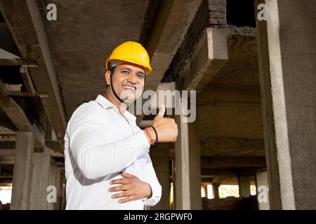 Porträt eines glücklichen jungen, gutaussehenden indischen Bauingenieurs oder Architekten, der ein weißes Hemd und einen gelben Schutzhelm trägt und beim Konstrukt Daumen nach oben zeigt Stockfoto